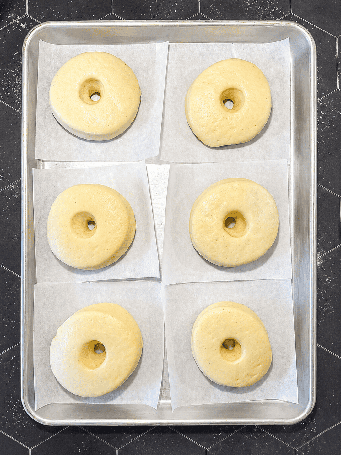Cut donuts rising on a sheet pan covered in parchment paper piece pieces.