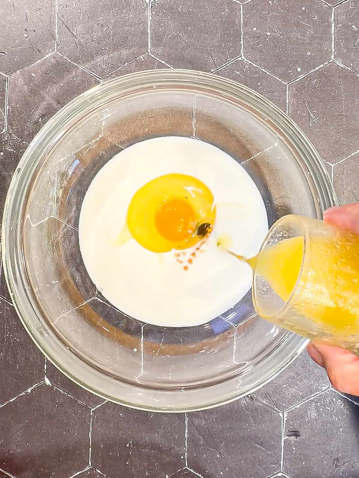 Pouring melted and cooled butter into wet ingredients in a glass bowl to make fritter batter.
